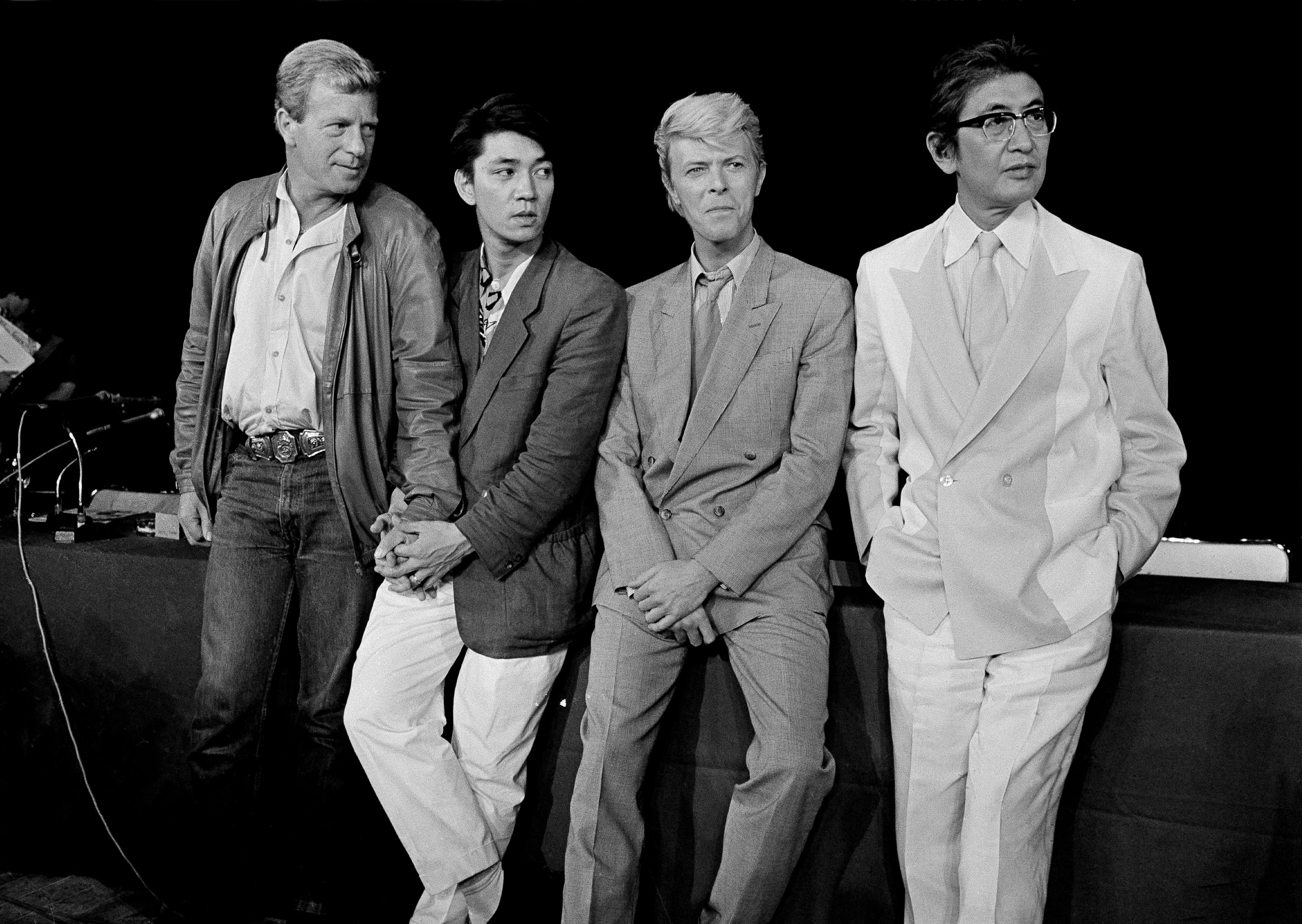 坂本龙一（左二）在电影《圣诞快乐，劳伦斯先生》中出演过一个角色，并负责配乐。 和他一起的是（左起）制片人杰克·托马斯、主演大卫·鲍伊和导演大岛渚。